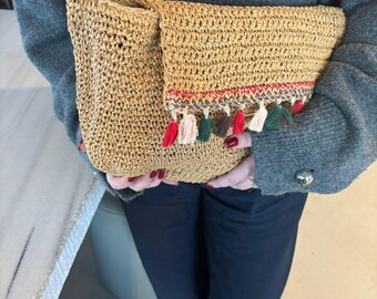May include: A woven raffia clutch in a natural tan color. The clutch features a fold-over flap with a decorative trim of red, white, pink, green, and brown tassels. The bag is held by a person wearing a gray sweater and dark blue pants.