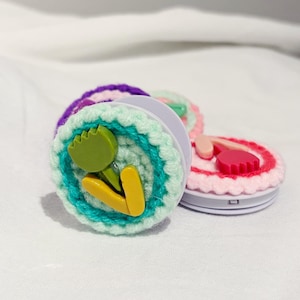 May include: A collection of crochet phone grips in various colors. One grip features a green tulip design with yellow accents, surrounded by teal and mint green crochet. Other grips have pink, purple, and red crochet details.