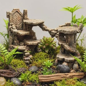 May include: A stone-coloured terrarium ornament featuring two tiered platforms connected by a bridge, surrounded by green moss, small ferns, succulents, smooth dark pebbles, and a piece of driftwood inside a glass enclosure.