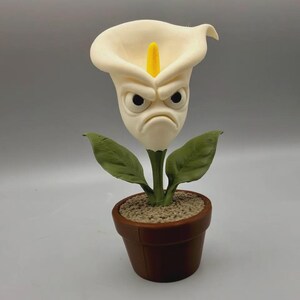 May include: A decorative figurine of a white calla lily flower with a grumpy facial expression, featuring a bright yellow spadix, two green leaves, and a textured brown pot filled with a granular material representing soil.