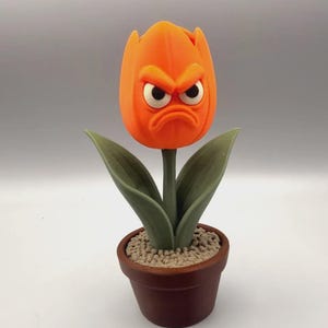 May include: A 3D printed orange tulip flower with a grumpy facial expression, featuring furrowed brows and a downturned mouth. The flower has two green leaves on a stem, planted in a small brown pot filled with textured light gray soil.