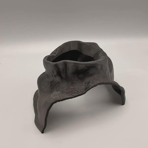May include: A matte black, three-dimensional object with an irregular, organic shape featuring a hollow center and a wide, arched opening at the base. The surface displays a fine, layered texture throughout, resembling topographical contour lines.