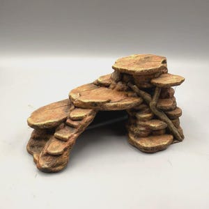 May include: A brown and tan terrarium ornament shaped like a tiered rock formation with a hollow cave entrance underneath. The structure features multiple flat, circular platforms resembling stone ledges and a textured branch-like accent.