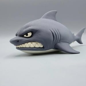 May include: A gray shark figurine with a textured surface, featuring a prominent dorsal fin, side fins, and a tail. The shark has a cartoonish expression with visible white teeth, a wide mouth, and narrow eyes set into its gray head.