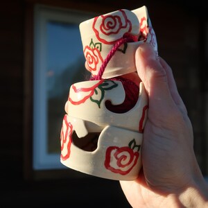 Può includere: Ciotole per filato in ceramica impilate, con base color crema e disegni di rose rosse. Un cordoncino rosa tiene insieme le ciotole. Le ciotole sono disposte verticalmente.