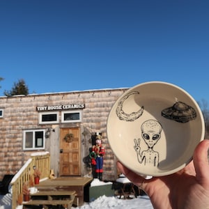 Peut inclure: Un bol en céramique crème avec des dessins noirs d'un croissant de lune, d'un OVNI et d'un extraterrestre faisant le signe de la paix. Le bol est tenu devant un bâtiment avec l'enseigne "Tiny House Ceramics".