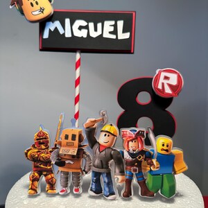 K&ouml;nnte beinhalten: Eine Kuchendekoration mit Roblox-Charakteren und dem Namen "MIGUEL". Der Aufsatz enth&auml;lt einen rot-wei&szlig; gestreiften Strohhalm, eine gro&szlig;e schwarze Zahl 8 und ein Roblox-Logo. Der Kuchen ist mit mehreren Roblox-Charakterfiguren dekoriert.
