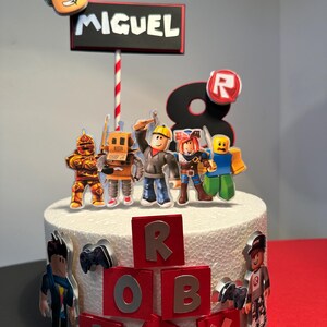 K&ouml;nnte beinhalten: Ein wei&szlig;er Kuchen, verziert mit Roblox-Figuren und einem Schild mit der Aufschrift "MIGUEL". Der Kuchen hat rote und silberne Akzente, darunter die Zahl "8" und das Wort "ROBLOX".