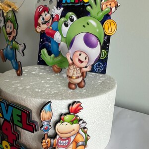 K&ouml;nnte beinhalten: Ein wei&szlig;er Kuchen, verziert mit bunten Cartoon-Charakter-Ausschnitten. Der Kuchen zeigt Charaktere wie Mario, Yoshi und Toad, mit dem Text "SUPER NIKO" und "LEVEL 4 LOCKED!". Der Kuchen ist mit einer Goldm&uuml;nze und einem Pinsel verziert.