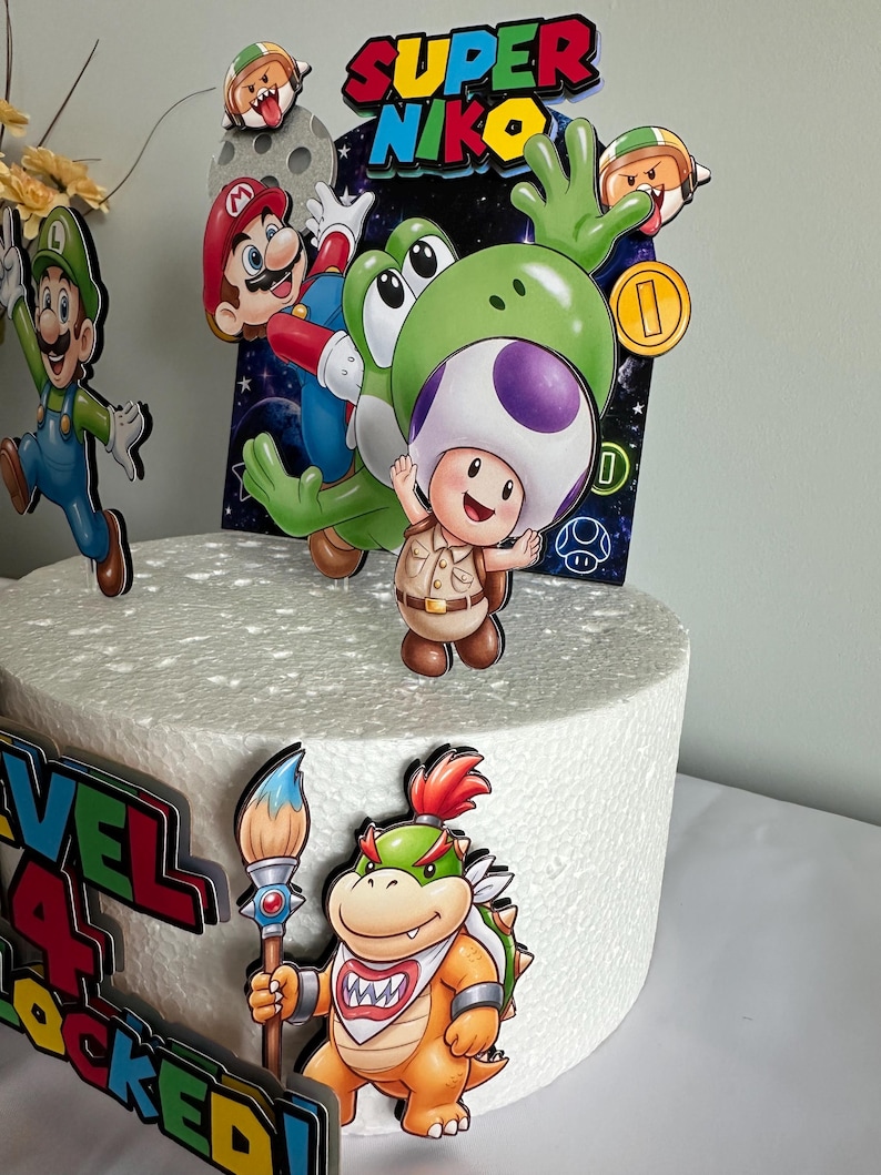 K&ouml;nnte beinhalten: Ein wei&szlig;er Kuchen, verziert mit bunten Cartoon-Charakter-Ausschnitten. Der Kuchen zeigt Charaktere wie Mario, Yoshi und Toad, mit dem Text "SUPER NIKO" und "LEVEL 4 LOCKED!". Der Kuchen ist mit einer Goldm&uuml;nze und einem Pinsel verziert.