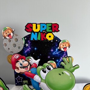 K&ouml;nnte beinhalten: Ein farbenfroher Tortenaufsatz mit Mario und Yoshi aus der Super Mario-Videospielreihe. Der Aufsatz enth&auml;lt den Text "SUPER NIKO" in Rot, Blau und Gelb. Zus&auml;tzliche Elemente sind ein Mond, M&uuml;nzen und andere Charaktere.