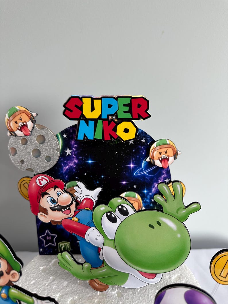 K&ouml;nnte beinhalten: Ein farbenfroher Tortenaufsatz mit Mario und Yoshi aus der Super Mario-Videospielreihe. Der Aufsatz enth&auml;lt den Text "SUPER NIKO" in Rot, Blau und Gelb. Zus&auml;tzliche Elemente sind ein Mond, M&uuml;nzen und andere Charaktere.