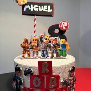 K&ouml;nnte beinhalten: Ein wei&szlig;er Kuchen, verziert mit Roblox-Figuren und dem Namen "MIGUEL" auf einem Schild. Der Kuchen zeigt rote und silberne Buchstaben, die "ROBLOX" buchstabieren, und eine gro&szlig;e "8". Verschiedene Roblox-Charaktere sind um den Kuchen herum platziert.