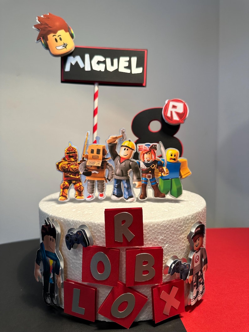 K&ouml;nnte beinhalten: Ein wei&szlig;er Kuchen, verziert mit Roblox-Figuren und dem Namen "MIGUEL" auf einem Schild. Der Kuchen zeigt rote und silberne Buchstaben, die "ROBLOX" buchstabieren, und eine gro&szlig;e "8". Verschiedene Roblox-Charaktere sind um den Kuchen herum platziert.