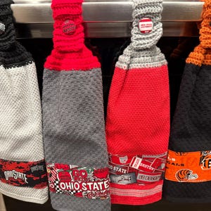May include: Four crocheted dish towels hanging on a rack. Each towel has a different team logo: Ohio State, Brutus Buckeye,  Go Bucks, and Bengals. The towels are gray, red, black, and orange.