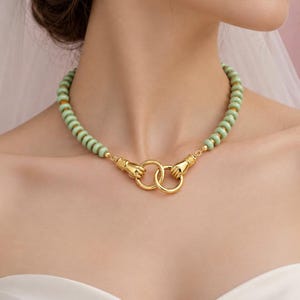 May include: A necklace featuring a strand of light green, disc-shaped beads. The ends of the bead strand connect to two gold-toned metal clasps shaped like hands. These hand-shaped clasps hold two interlocking gold-toned metal rings at the centre.