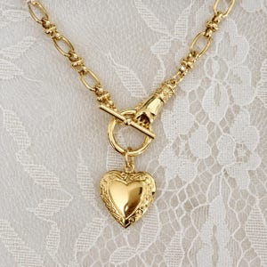 May include: A gold-toned necklace featuring a thick oval link chain and a toggle clasp shaped like a hand. A heart-shaped pendant with an ornate, raised border hangs from the circular ring of the toggle closure, creating a polished metallic look.