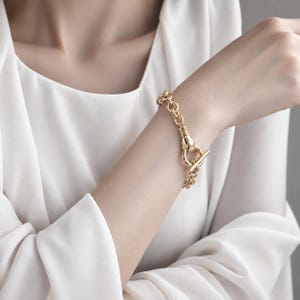 Könnte beinhalten: Ein goldfarbenes Gliederarmband mit einem markanten Knebelverschluss. Das Armband besteht aus polierten Metallgliedern und verfügt über einen T-Steg-Verschluss, der in einen kreisförmigen Ring greift und so ein durchgehendes Design bildet.