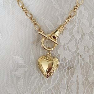 Puede incluir: Collar dorado con colgante de medallón en forma de corazón. El collar tiene un cierre de palanca y una cadena con eslabones decorativos. El medallón tiene un diseño ornamental en relieve. El fondo es una tela blanca y de encaje.