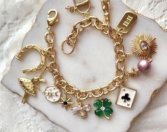 Op de afbeelding: Een goudkleurige schakelarmband met diverse bedels, waaronder een hoefijzer, een ballerina, een zon- en maanschijf, een ster, een bij, een groen klavertjevier, een klaveren aas speelkaart, een parel, een zon-oog en een hangslot.