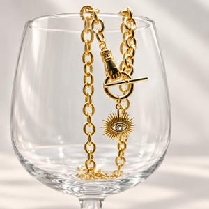 May include: A gold-toned chain necklace featuring a toggle clasp shaped like a hand holding a ring. A sunburst-style pendant with a central clear stone shaped like an eye hangs from the toggle ring. The necklace is draped inside a clear glass.