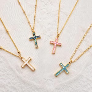 Könnte beinhalten: Vier goldfarbene Halsketten mit Kreuzanhängern. Jedes Kreuz ist mit kleinen, quadratischen Plättchen in verschiedenen Farben eingelegt: eines mit schillerndem Blau und Grün, eines in Hellrosa, eines in Weiß und eines in hellem Blau.
