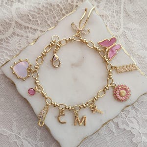 Könnte beinhalten: Goldfarbenes Charm-Armband mit verschiedenen Anhängern, darunter ein rosa Schmetterling, eine Gänseblümchen, ein Herz und die Buchstaben "C" und "M". Das Armband hat einen Karabinerverschluss und wird auf einer weißen Marmoroberfläche präsentiert.