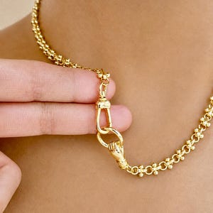 Può includere: Una collana a catena color oro con un design a maglie testurizzate. La collana include un meccanismo di chiusura prominente a forma di mano che regge un anello circolare, collegato a una maglia secondaria a forma di U.