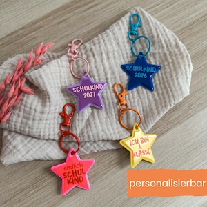 Könnte beinhalten: Eine Auswahl an sternförmigen Schlüsselanhängern in verschiedenen Farben, darunter Pink, Lila, Blau und Gelb. Jeder Schlüsselanhänger trägt einen anderen Text, wie "SCHULKIND 2026" und "ICH BIN 1. KLASSE". Die Schlüsselanhänger haben Metallverschlüsse.