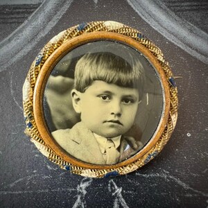 May include: Vintage circular brooch featuring a sepia-toned portrait of a young child. The portrait is framed by a wooden border, surrounded by a gold-toned, braided trim with blue accents. The brooch has a pin on the back.