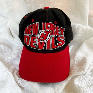 Può includere: Un cappellino da baseball nero con visiera rossa, caratterizzato dalla scritta ricamata NEW JERSEY DEVILS in rosso con bordi bianchi e un logo centrale a forma di diavolo rosso. Il cappellino presenta una corona strutturata e una visiera curva.