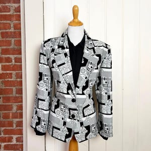 May include: A monochrome patterned blazer with a geometric design, displayed on a wooden mannequin. The blazer features a notched lapel, two buttons, and long sleeves. A black collared shirt is worn underneath.