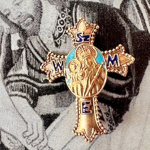 May include: A gold-colored cross pendant with a detailed design. The cross features a central oval with a religious image, surrounded by blue enamel. The arms of the cross have the letters W, M, S, and E. The cross is likely a religious item.