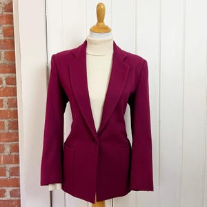 May include: A deep magenta blazer with a classic tailored design. The blazer features a single-button closure, notched lapels, and two front pockets. The garment is displayed on a mannequin, paired with a cream-colored turtleneck.