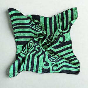 Puede incluir: Un pañuelo cuadrado de seda con un patrón geométrico. El pañuelo presenta rayas alternas verdes y negras, con un diseño repetitivo de formas entrelazadas. El pañuelo está doblado y drapeado, mostrando su textura y diseño.