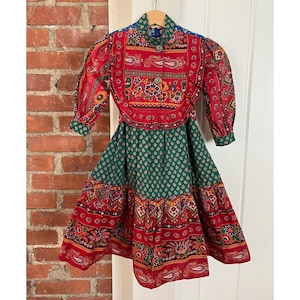 Puede incluir: Un vestido vintage con un diseño vibrante y estampado. El vestido presenta un corpiño rojo con motivos florales y paisley, una falda verde con un patrón repetido y una falda roja escalonada. Las mangas son abullonadas y el vestido está colgado en una percha de madera.