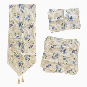 Könnte beinhalten: Ein beigefarbener Tischläufer und ein passendes Servietten-Set. Der Stoff ist mit blauen und violetten Blumenmustern versehen. Der Tischläufer hat Quasten an den Enden. Die Servietten sind quadratisch und gestapelt.