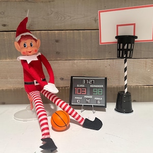 May include: A red and white elf doll in a basketball-themed scene. The elf is wearing a red outfit with white accents and striped legs, holding an orange basketball. A miniature scoreboard displays scores, and a basketball hoop is in the background.