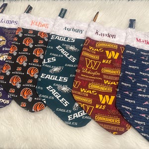 May include: Five Christmas stockings with different NFL team logos. The stockings are purple, black, green, maroon, and blue. The names James, Ethan, Anthony, Kavden, and Malkem are written on the stockings.