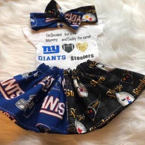 Puede incluir: Un conjunto de bebé con un body blanco, una diadema azul y blanca y una falda negra y azul. El body tiene el texto "I'm Divided but love Mommy and Daddy the same!" y los logotipos de los New York Giants y los Pittsburgh Steelers. La falda está dividida en dos, con un lado con el logotipo de los New York Giants y el otro lado con el logotipo de los Pittsburgh Steelers.