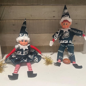 Peut inclure: Deux poupées lutins vêtues de tissu vert foncé avec le logo et le texte des Philadelphia Eagles. L'un porte une robe et tient des pompons dorés, l'autre porte un haut et un pantalon et est assis près d'un petit ballon de football marron.