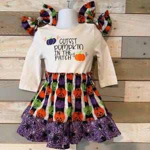 Könnte beinhalten: Ein weißes Langarmshirt mit dem Text CUTEST PUMPKIN IN THE PATCH und Kürbisgrafiken, kombiniert mit einem Rock mit orangefarbenen, grünen und violetten Kürbissen auf schwarz-weißen Streifen sowie einem violetten Rüschensaum mit Spinnennetz.