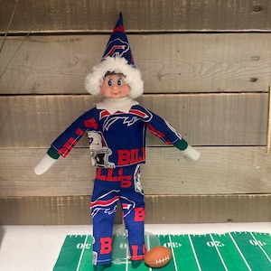 May include: An elf doll dressed in a blue and red Buffalo Bills football uniform, including a pointed hat and white collar. A small American football sits on a green field-patterned surface.