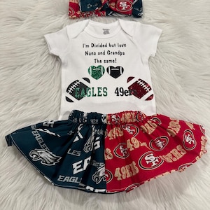 May include: A white baby onesie with the text "I'm Divided but love Nana and Grandpa The same!" and two footballs with the words "Eagles" and "49ers". The baby is wearing a green and red skirt with the logos of the Philadelphia Eagles and the San Francisco 49ers.