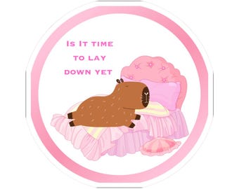 Peut inclure: Autocollant rond avec une bordure rose. Il représente un capybara de dessin animé sur un lit rose avec une tête de lit touffetée et une literie à volants. Le texte "IS IT TIME TO LAY DOWN YET" est écrit en rose. Design mignon.