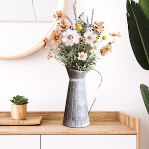 May include: A galvanized metal pitcher filled with a vibrant arrangement of flowers. The bouquet includes white, peach, and yellow blooms, with green foliage. The pitcher sits on a wooden surface, with a small succulent in a wooden pot nearby.