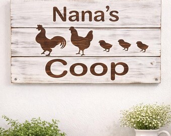 May include: A rustic wooden sign with the words "Nana's Coop" and silhouettes of a rooster, hen, and chicks. The sign is white with brown lettering and images, and is hanging by a rope.