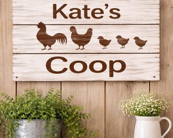 May include: A rectangular white-washed wooden sign hanging by a rope, featuring the brown text Kate's Coop and silhouettes of a rooster, a hen, and three chicks. The sign is constructed from three horizontal planks with visible wood grain.