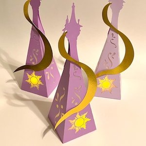 Peut inclure: Trois boîtes de faveur de fête violettes en forme de cône avec des accents dorés. Chaque boîte présente un motif de soleil jaune et des volutes dorées. Une découpe dorée en forme de ruban est attachée à chaque boîte.