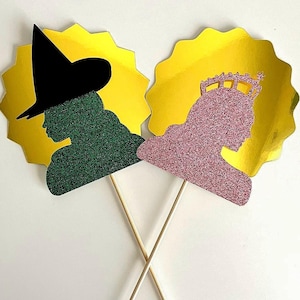 May include: Two gold glitter photo booth props with stick handles. One prop is a green silhouette of a witch wearing a black hat. The other prop is a pink silhouette of a queen wearing a crown.